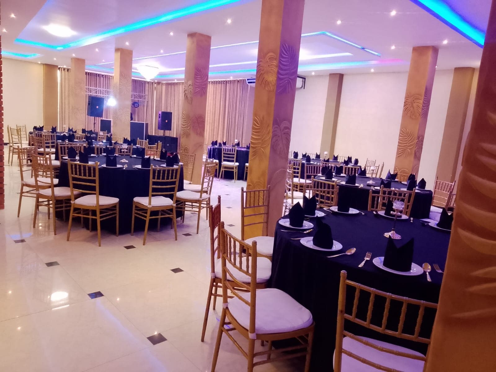 The reception hall set with round tables, black linens and gold chairs under coloured cove lighting
